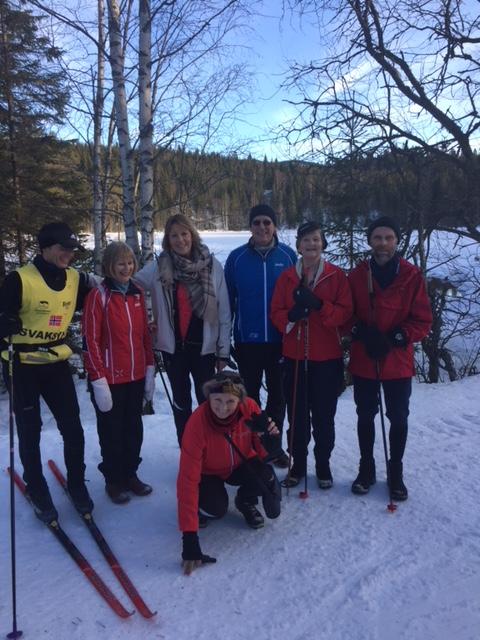 Gruppebildet viser 7 personer, en med ski på bena. I bakgrunnen ses et snødekt vann med skog på andre siden.