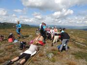 Bildet viser gruppen liggende hensllengt rundt omkring oppe på fjellet. Noen sitter ved et utplasert bord. Det er nydelig vær og helt åpent terreng. I bakgrunnen ser en så vidt Vangsjøen.