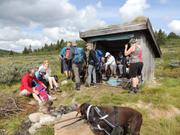 Bildet viser gruppen samlet for lunsj ved og i en gapahuk bygd av treverk. I forgrunnen står hunden Taro, som var med på turen